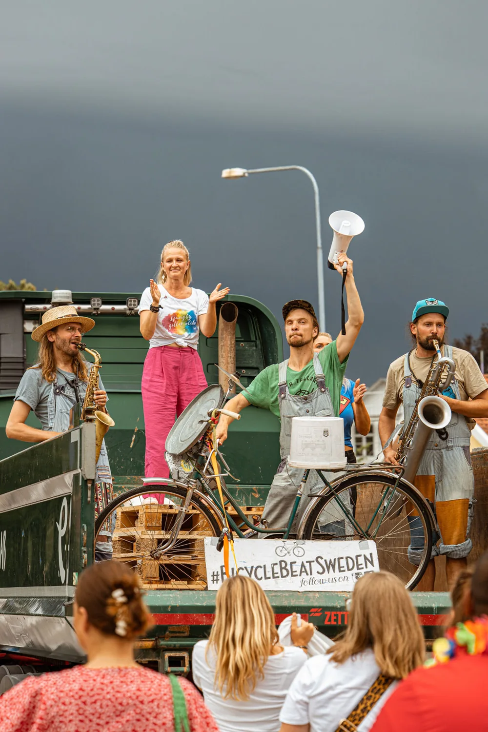 Bandet spelar uppe på ett flak utomhus med megafon, saxofon och cykel framför publik.